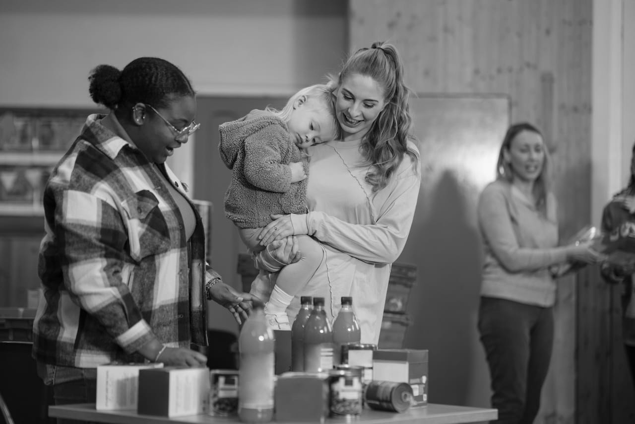 Two people look at a table with diluting juice, cans and tea. One is holding a forlorn looking small child and the other is looking enthusiastically at the juice.