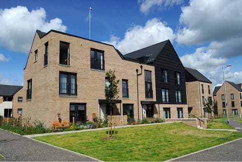 A block of flats with an angular geometry. They feature a small tended garden and grass in front of them.