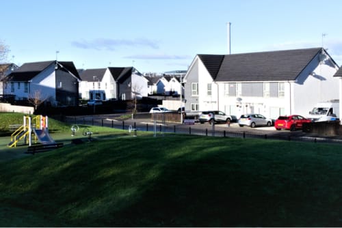 A neighbourhood of white terraced houses. A playpark is shown in the foreground and there are several cars parked in the driveways.
